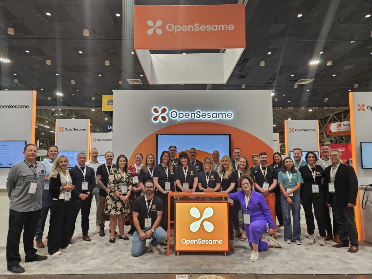 A large group of OpenSesame employees poses for a group photo in front of their expo booth.