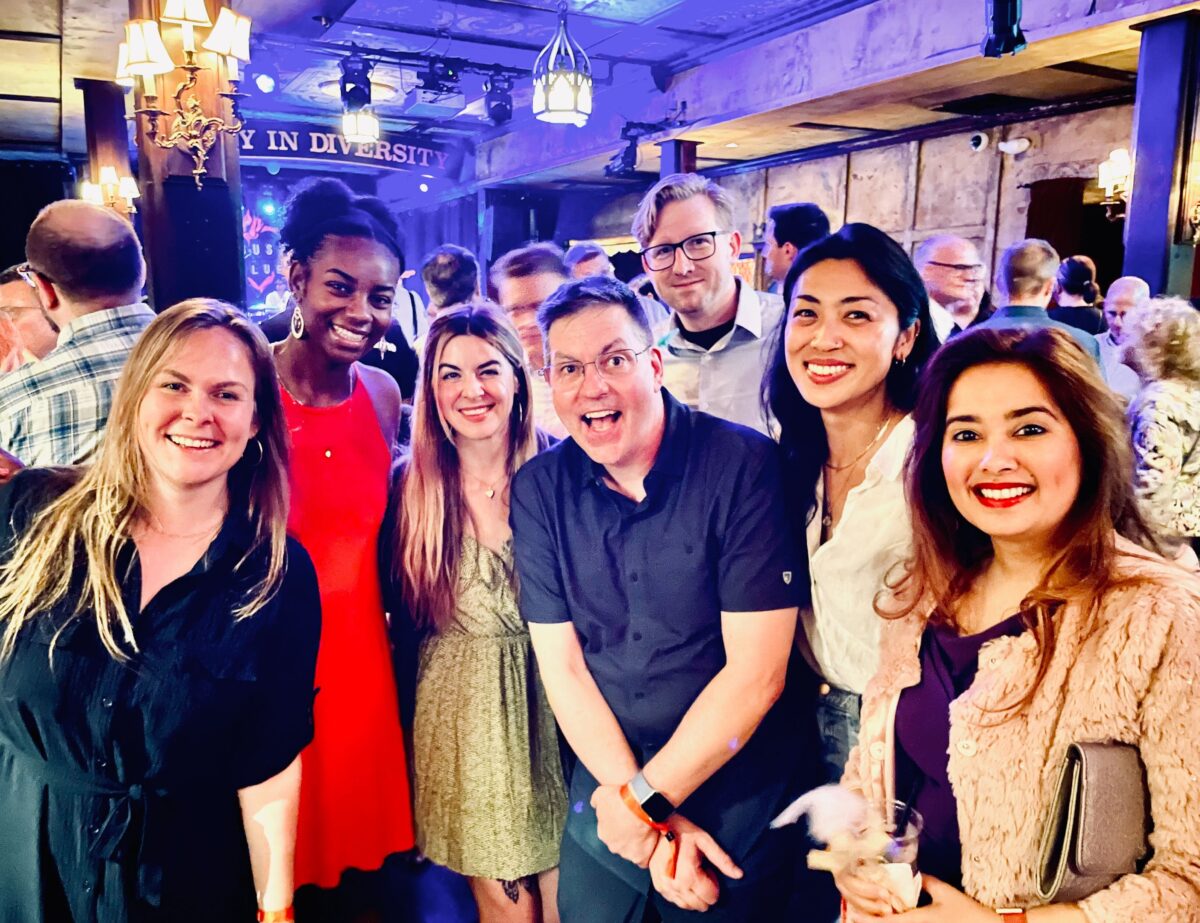 A diverse group of seven people is gathered together in a lively indoor setting, smiling for the camera. The background is dimly lit with warm lighting and chandeliers, and the phrase "Unity in Diversity" is visible on a sign behind them. The individuals are dressed in a mix of casual and semi-formal attire, with one woman wearing a bright red dress standing out prominently. The atmosphere appears to be cheerful and festive, suggesting a social event or gathering.