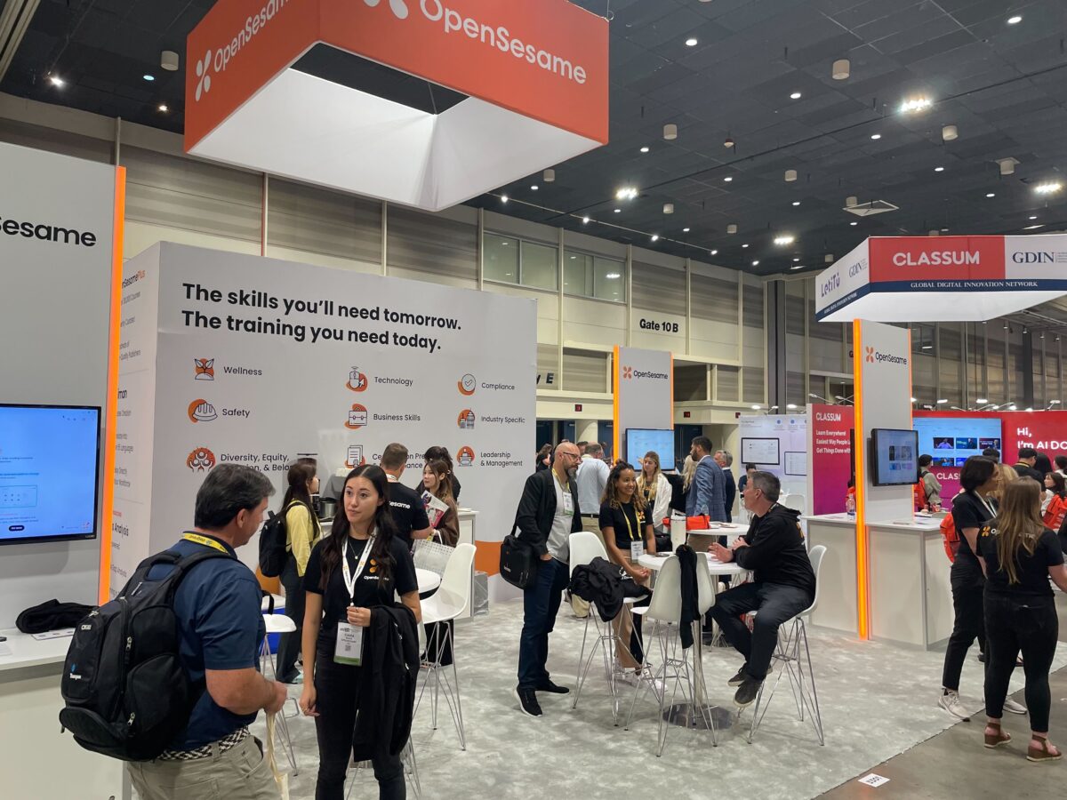 A bustling trade show or conference exhibit hall with an OpenSesame booth prominently displayed. The booth features a large sign with the company's name and the tagline "The skills you'll need tomorrow. The training you need today." Various icons and text highlight different training areas such as Wellness, Technology, Safety, Compliance, and Business Skills. Several attendees are engaged in conversations with representatives at the booth. Some people are sitting at high tables, while others stand and chat. The environment is professional and energetic, with other booths and attendees visible in the background.